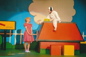 Scene from the show with Snoopy sitting on his kennel with Sally standing in front of him