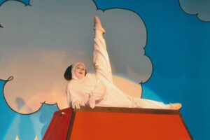 Snoopy in a dance routine, lying on his side on top his kennel with his leg pointed at the sky