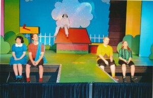 Cast members sitting on the edge of the stage while Snoopy sits on his kennel