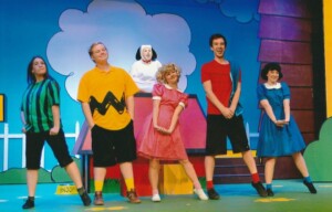 The cast singing and dancing in front of Snoopy sitting on his kennel